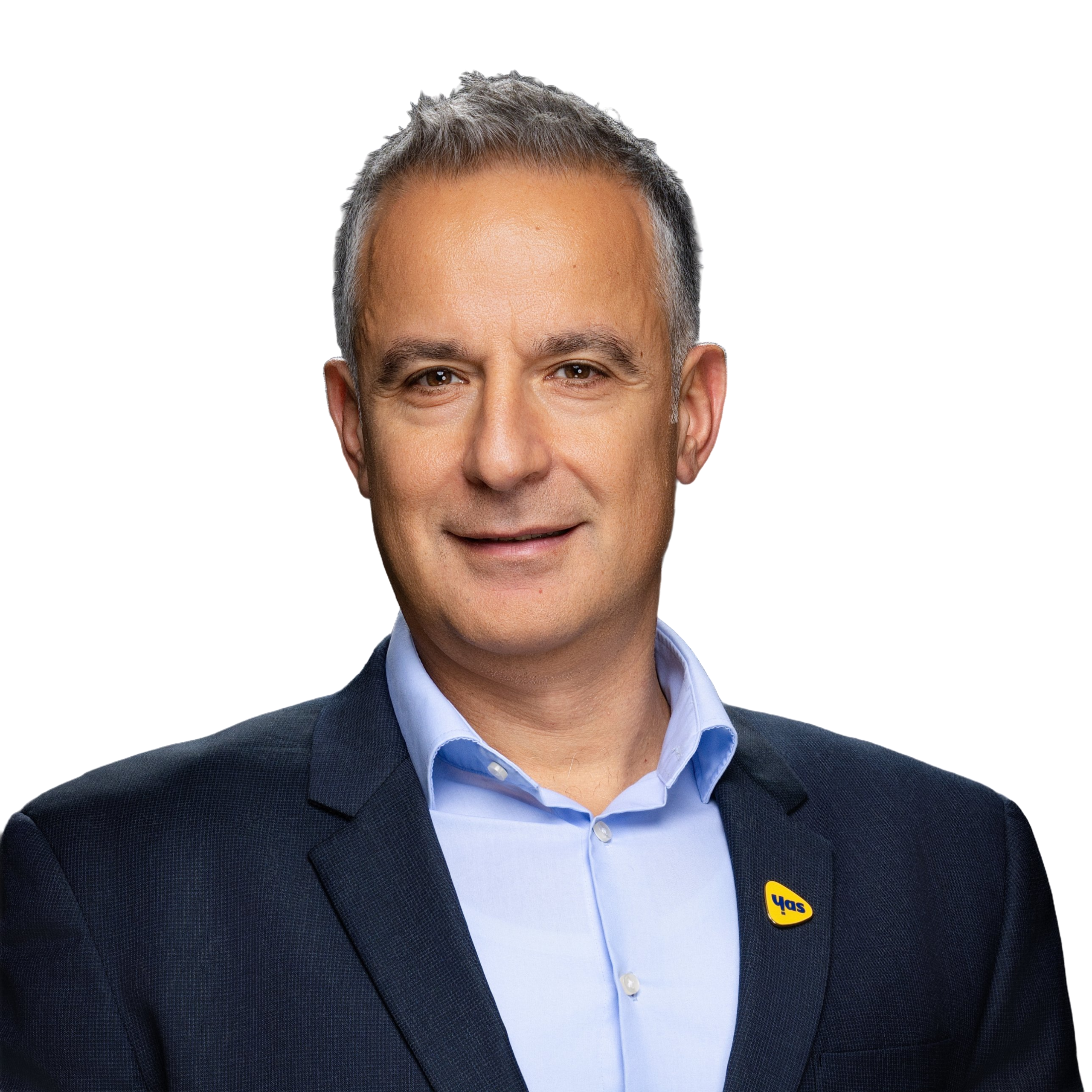 A middle-aged man with short, greying hair smiles against a light grey background. He is wearing a dark blue suit jacket over a light blue button-down shirt, with a small, yellow triangular lapel pin featuring the text “yas” attached to his jacket.