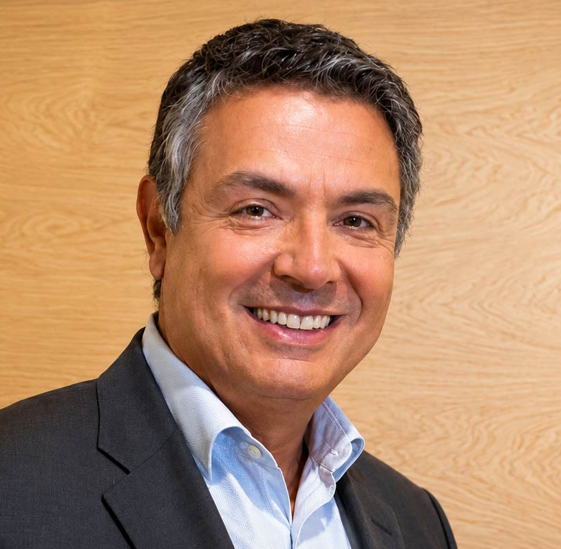 A middle-aged man with salt-and-pepper hair smiles at the camera. He is wearing a dark grey suit jacket over a light blue button-down shirt against a plain, light wood-grain background.