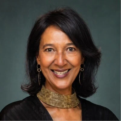 A smiling woman with shoulder-length dark hair wears a textured gold choker necklace and gold hoop earrings. She is dressed in a black top against a dark, muted green background.