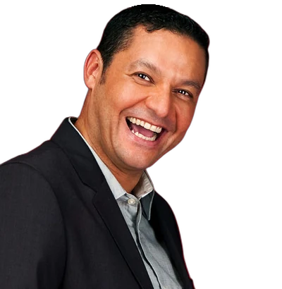 A smiling middle-aged man with short dark hair and a tanned complexion looks towards the camera. He is wearing a dark suit jacket over a light-coloured button-down shirt against a plain white background.