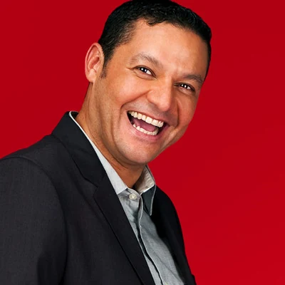 A smiling man with short dark hair wears a grey button-down shirt and a black blazer. He is positioned against a vibrant, solid red background, looking towards the camera with an open-mouthed expression of joy.