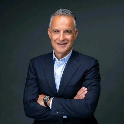 A middle-aged man with short, greying hair smiles directly at the camera with his arms crossed. He is wearing a dark navy blazer over a light blue collared shirt against a plain, dark grey background.
